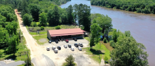 Aerial view of a riverside building with parked cars, surrounded by trees and a flowing river.