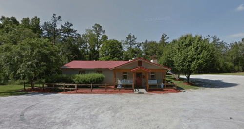 A single-story building with a red roof, surrounded by trees and a gravel parking area.