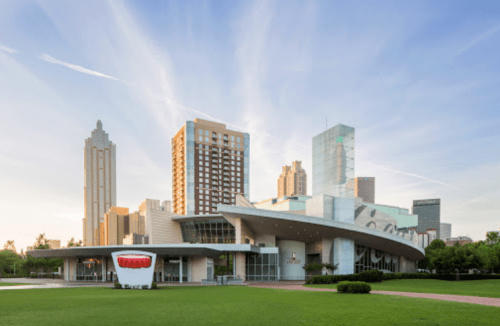 Modern building with a curved roof, surrounded by green grass and city skyscrapers under a clear blue sky.