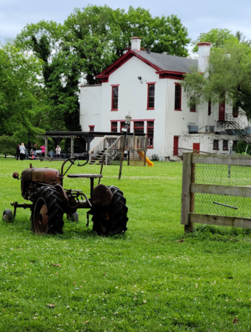 An old tractor sits in a grassy field in front of a historic building with red accents and trees in the background.
