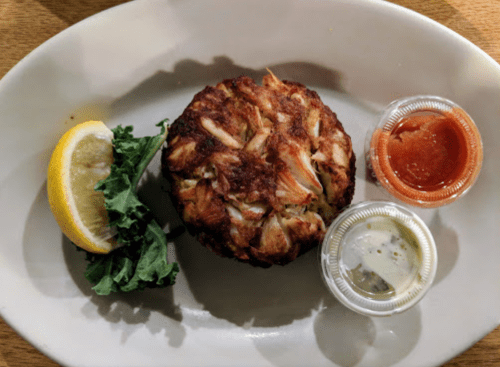 A crab cake on a plate, accompanied by lemon, kale, and two dipping sauces.