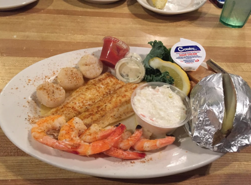 A seafood platter featuring grilled fish, shrimp, scallops, coleslaw, a baked potato, and lemon garnish.