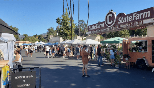 A bustling outdoor market with tents, vendors, and people shopping near a stadium entrance. Palm trees in the background.