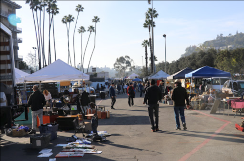 A bustling outdoor market with vendors under tents, people browsing, and palm trees in the background.