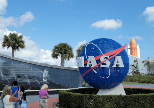 Visitors take photos near a large NASA logo and a space shuttle in the background, with palm trees and blue skies.