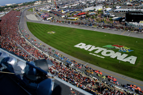 Aerial view of a crowded Daytona racetrack with cars racing and fans in the stands during a NASCAR event.