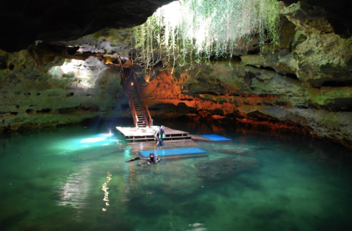 A serene cave with turquoise water, a wooden platform, and hanging greenery, illuminated by natural light from above.