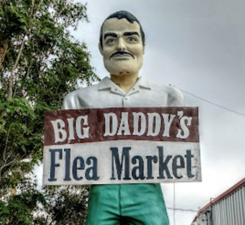 A large statue of a man in green pants and a white shirt stands in front of a sign for "Big Daddy's Flea Market."