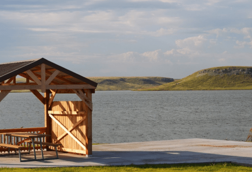 A wooden gazebo by a calm lake, surrounded by green hills under a partly cloudy sky.