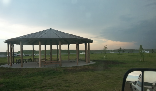 A gazebo with wooden pillars in a grassy area, under a cloudy sky with a hint of sunset in the background.