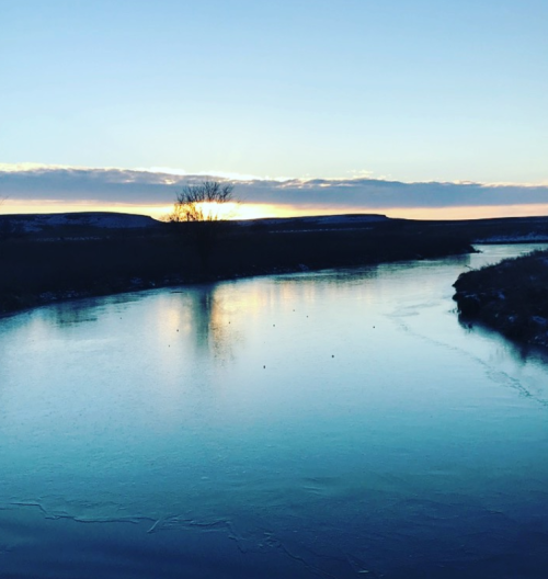 A serene river at sunset, reflecting the sky with a silhouette of trees and hills in the background.