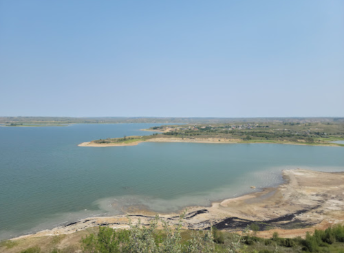 A serene landscape featuring a calm lake surrounded by land and distant hills under a clear blue sky.