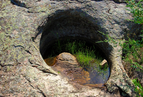 A natural rock formation with a circular depression filled with water and surrounded by green grass.