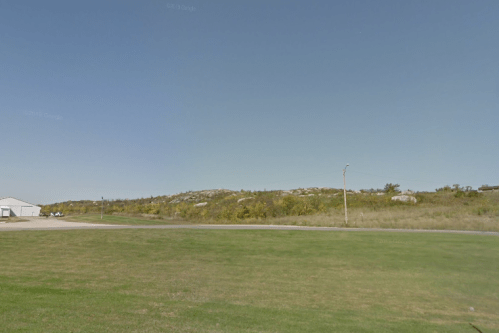 A grassy area with a clear blue sky, featuring a distant hill and a few trees, near a road and a building.