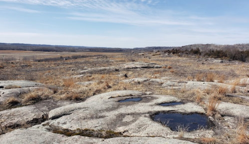 A rocky landscape with patches of grass and small puddles, under a clear blue sky and distant hills.