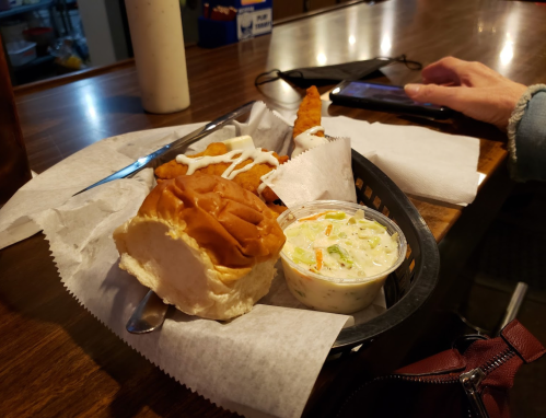 A plate with a sandwich, coleslaw, and a fried item, served on a wooden table with a phone nearby.
