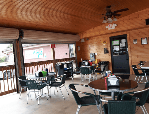 A cozy indoor dining area with wooden walls, round tables, and green chairs, featuring a ceiling fan and exit sign.