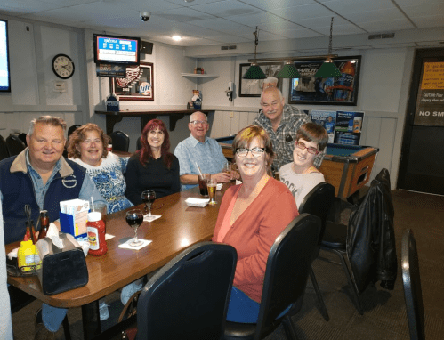 A group of seven people sitting around a table in a casual setting, smiling and enjoying drinks.