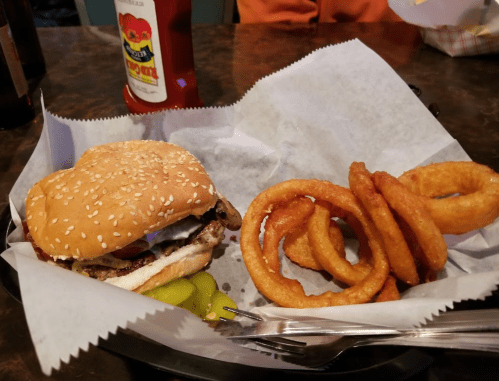 A sesame seed burger with pickles and onion rings on a paper-lined plate, accompanied by ketchup.