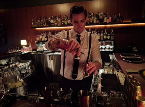 A bartender in a black tie and suspenders pours a cocktail at a stylish bar with shelves of liquor in the background.