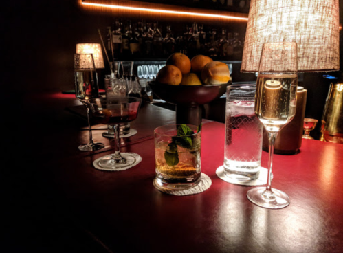 A dimly lit bar scene featuring various cocktails and a bowl of fruit on the counter.