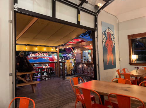 A cozy restaurant interior with wooden tables, orange chairs, and a vibrant mural, featuring a burger counter in the background.