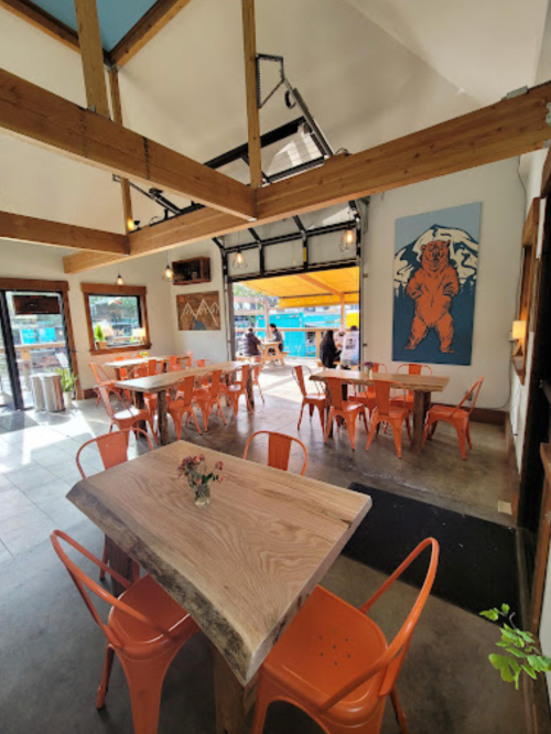 Bright interior of a café with wooden tables, orange chairs, and a bear mural on the wall, featuring large windows.