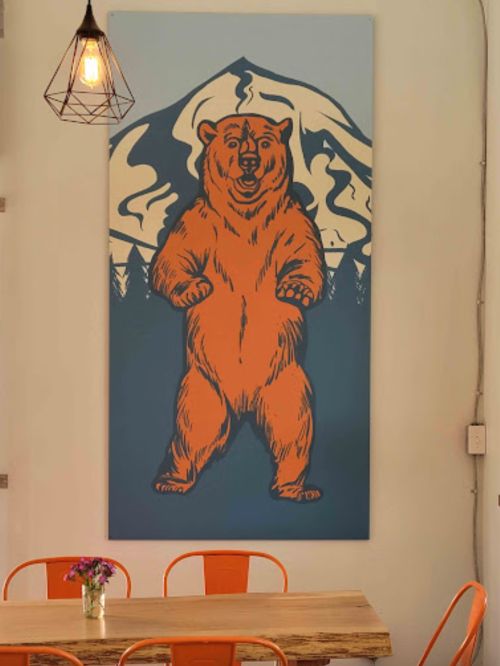 A large bear mural stands on a wall above a wooden table with orange chairs and a small vase of flowers.