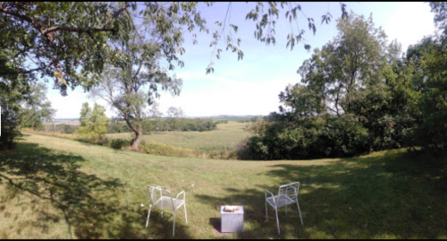 A panoramic view of a grassy landscape with two white chairs and a small table, surrounded by trees and open fields.
