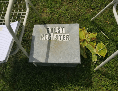A metal box labeled "GUEST REGISTER" sits on grass, next to a plant and two white chairs.