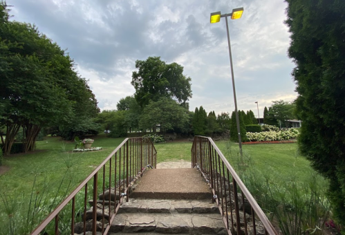 A stone pathway leads to a grassy area, flanked by trees and flowers, under a cloudy sky. A lamp stands nearby.