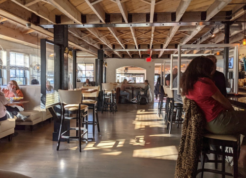 A spacious restaurant interior with wooden beams, booths, and patrons dining at tables and the bar. Natural light filters in.