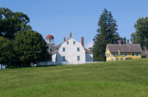 A white historic building and a yellow house sit on a grassy hill, surrounded by trees under a clear blue sky.