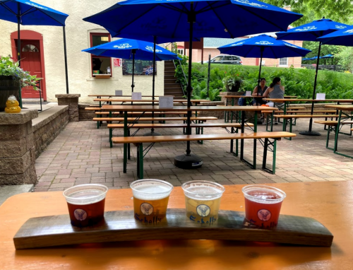 A wooden flight with three beer samples sits on a table in an outdoor seating area with blue umbrellas and empty benches.