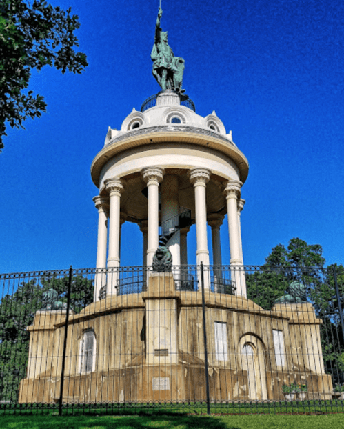 A tall, circular monument with columns and a statue on top, surrounded by a fence and greenery under a clear blue sky.