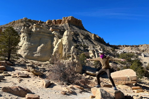 A person stands on a rock in a desert landscape, striking a pose with cliffs and blue sky in the background.