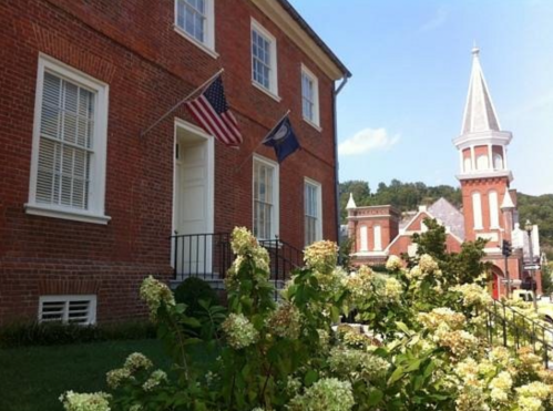 Historic brick building with American and state flags, surrounded by blooming flowers and a church steeple in the background.