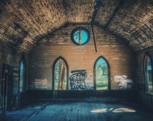 Interior of an abandoned building with wooden walls, arched windows, and graffiti on one wall. Sunlight filters in.