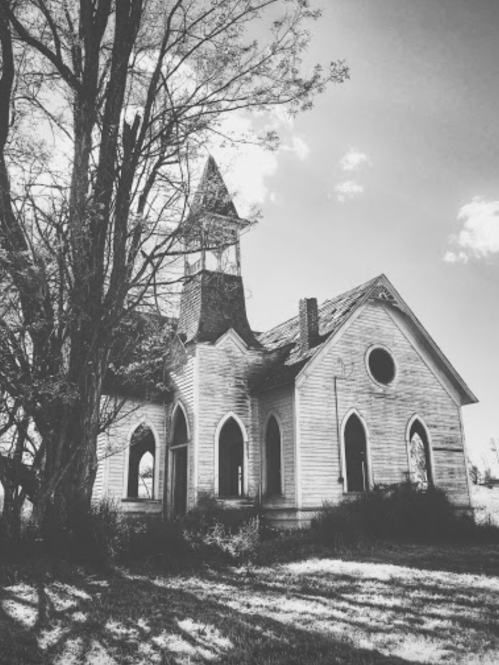 A weathered, abandoned church with a tall steeple, surrounded by trees, captured in black and white.