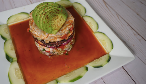 A colorful layered dish with avocado on top, surrounded by cucumber slices and drizzled with sauce on a white plate.