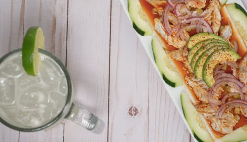 A platter of shrimp topped with onions and avocado slices, alongside a glass of iced drink with a lime wedge.