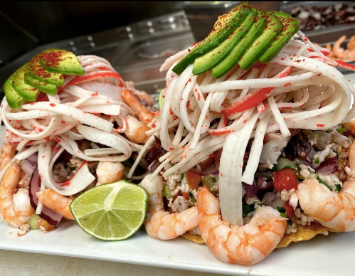 Two shrimp tacos topped with shredded vegetables, avocado slices, and lime wedges on a white plate.