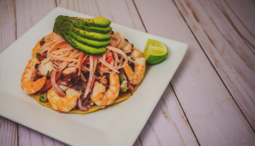 A shrimp taco topped with avocado slices, pickled onions, and a lime wedge on a white plate.