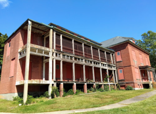Abandoned brick building with a weathered porch and overgrown grass under a clear blue sky.
