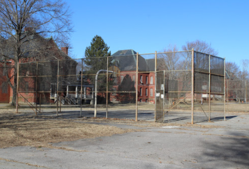 Abandoned basketball court with a chain-link fence, surrounded by overgrown grass and a derelict building in the background.