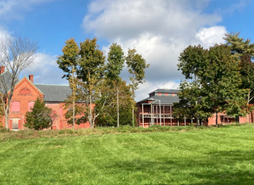 Historic red brick buildings surrounded by trees and a grassy field under a partly cloudy sky.