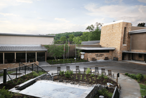 A modern lodge building with stone accents, surrounded by greenery and a small water feature in the foreground.
