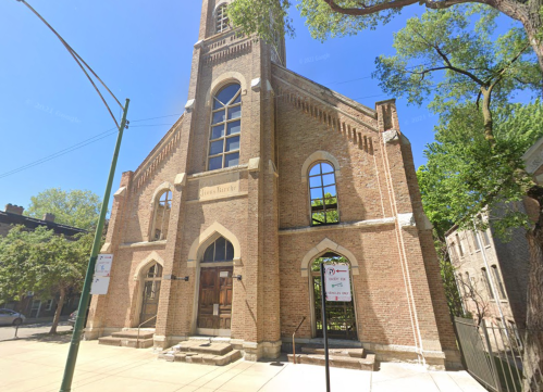 Historic brick church with tall windows and a steeple, surrounded by trees and street signs.