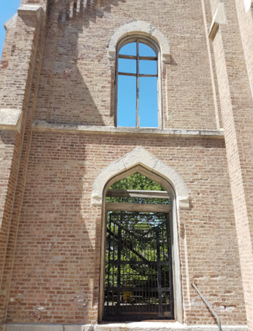 A brick wall of a church with a large arched doorway and a window above, framed by trees and blue sky.