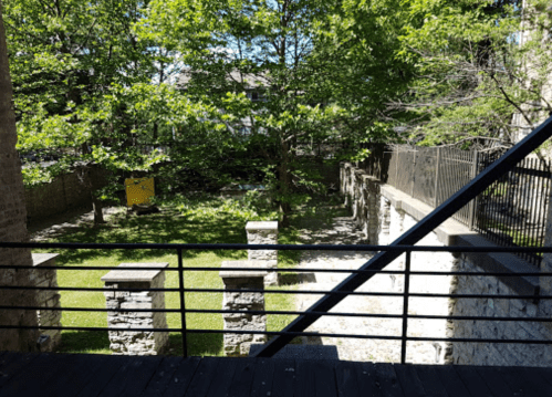 A view of a green garden with trees and stone walls, seen from a balcony with a metal railing.